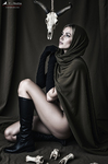 Oriana sentada con capa negra y capucha bajo un cráneo de animal, rodeada de huesos y objetos rituales.